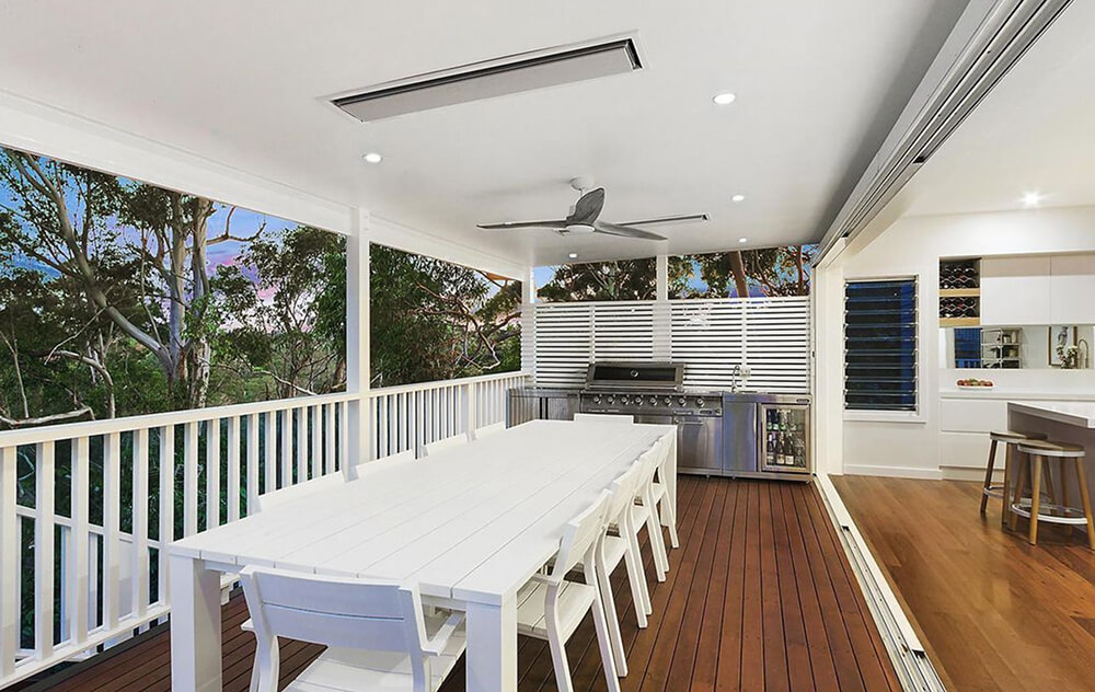 Sleek, covered outdoor dining area with a long white table and chairs, featuring a stainless steel barbecue and kitchen setup, ceiling fan, and seamless transition to the indoor kitchen, surrounded by lush greenery.