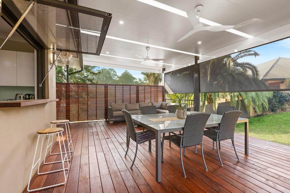 Contemporary alfresco dining area with a glass-top table and wicker chairs, featuring retractable blinds, ceiling fans, and an outdoor bar counter, seamlessly connected to the kitchen, perfect for relaxed outdoor entertaining.