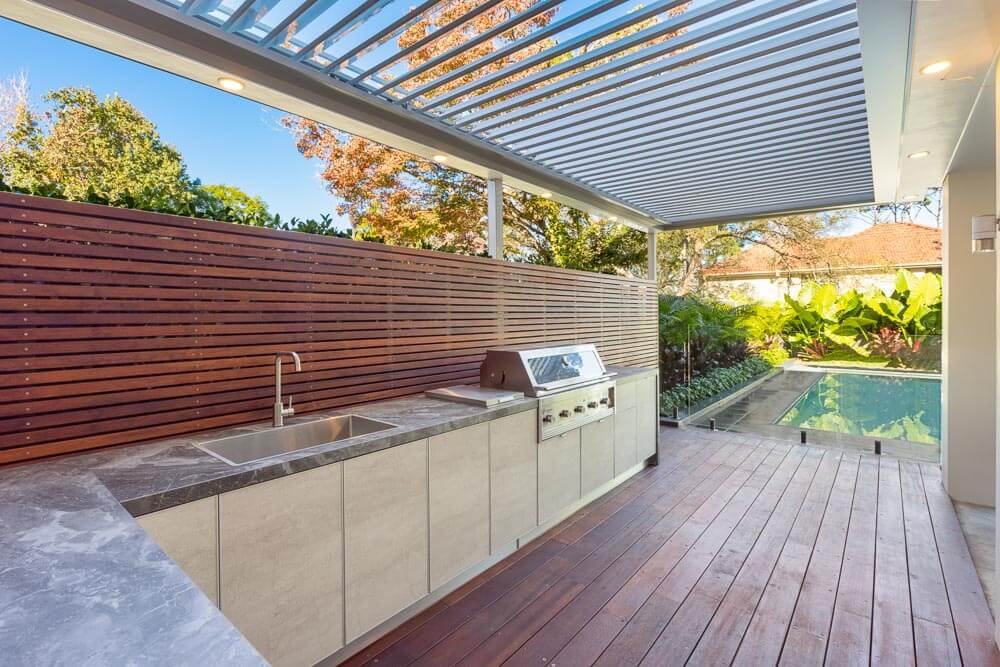 Modern outdoor kitchen with marble countertops, stainless steel appliances, and a louvered roof, adjacent to a wooden deck overlooking a pool, creating a stylish and functional space for outdoor cooking and relaxation.