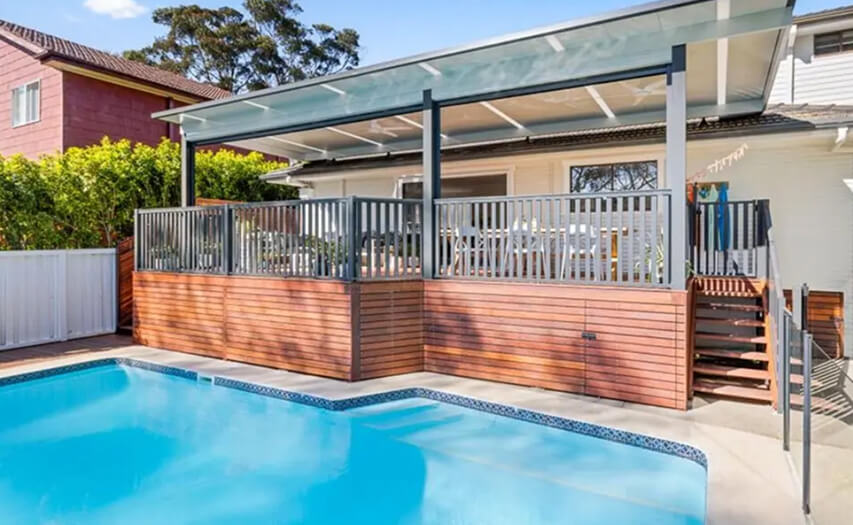 Custom elevated timber deck and integrated alfresco patio cover built close to a property boundary line by Emanate and Co, featuring modern poolside safety balustrading and an outdoor room extension.