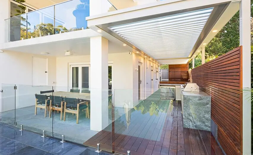 Custom outdoor kitchen and integrated louvred patio room built along a property boundary line by Emanate and Co, featuring stone countertops, glass pool balustrading, and a timber privacy screen.