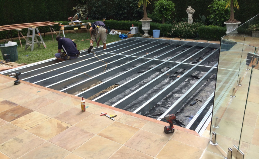 Mid-construction view of an engineered low-to-ground galvanized steel deck subframe by Emanate and Co, featuring two builders actively installing timber decking boards next to a pool zone.
