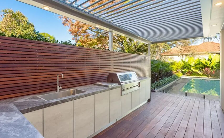 Custom outdoor kitchen island and integrated louvred patio roof extension by Emanate and Co, featuring premium timber deck flooring, a privacy slat wall, and an adjacent swimming pool.