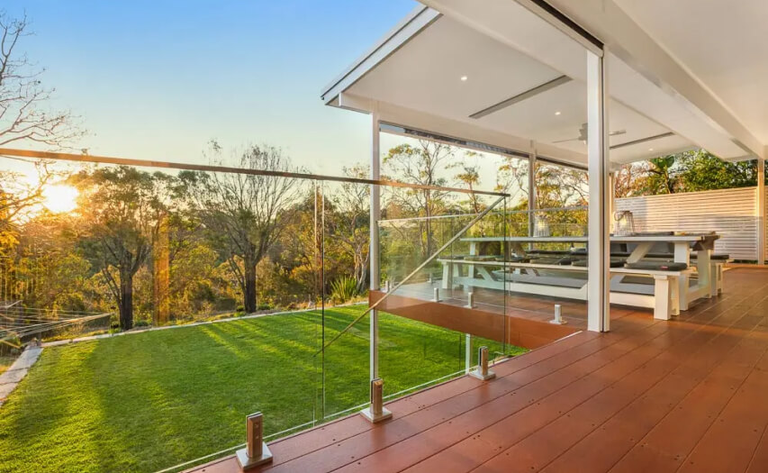 Luxury elevated timber deck and custom outdoor room extension by Emanate and Co, featuring premium glass balustrades, an integrated outdoor dining zone, and sleek flyover patio ceiling fixtures at sunset.