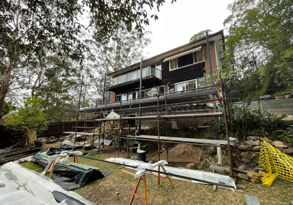 Multi-level residential deck extension under construction by Emanate and Co, showing subfloor steel framing, structural steel posts, and safety scaffolding on a sloping block.