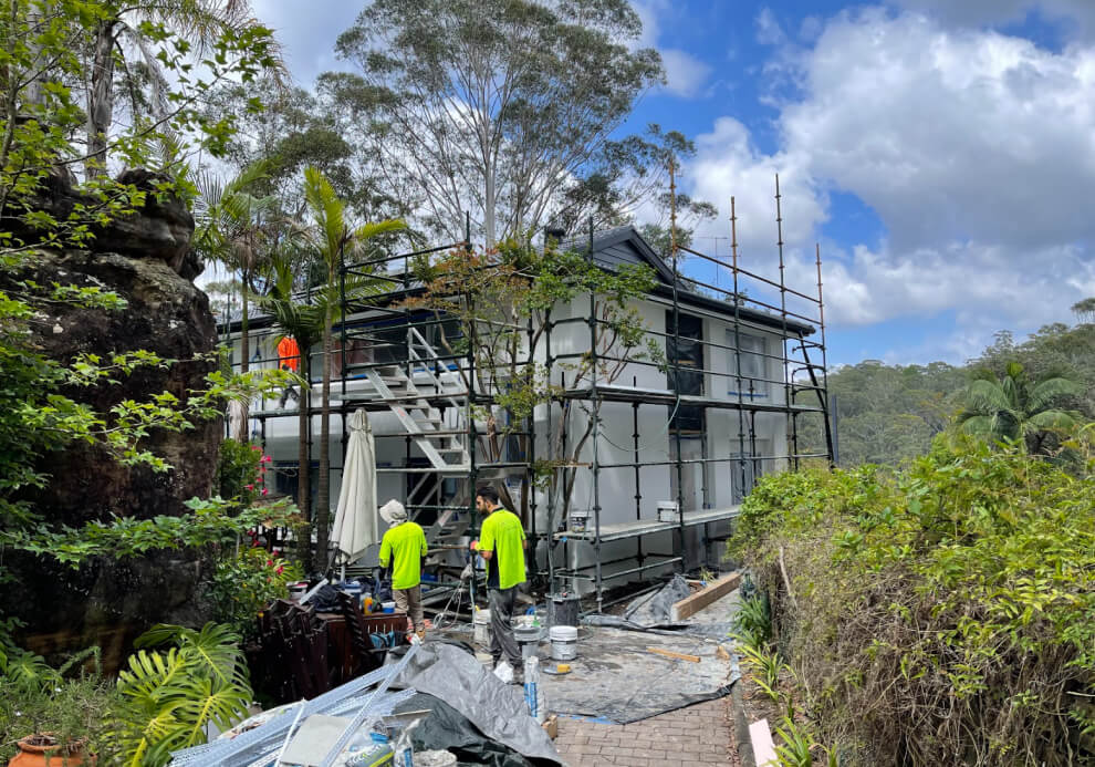Multi-story residential home extension under construction by Emanate and Co, featuring professional site scaffolding, structural steel framing, and builders on a challenging sloping site.