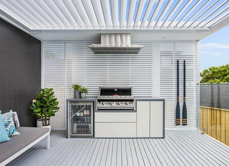 Modern alfresco outdoor kitchen with built-in stainless steel BBQ, glass-door beverage fridge, stainless rangehood, and opening louvre roof in Balgowlah by Emanate and Co.