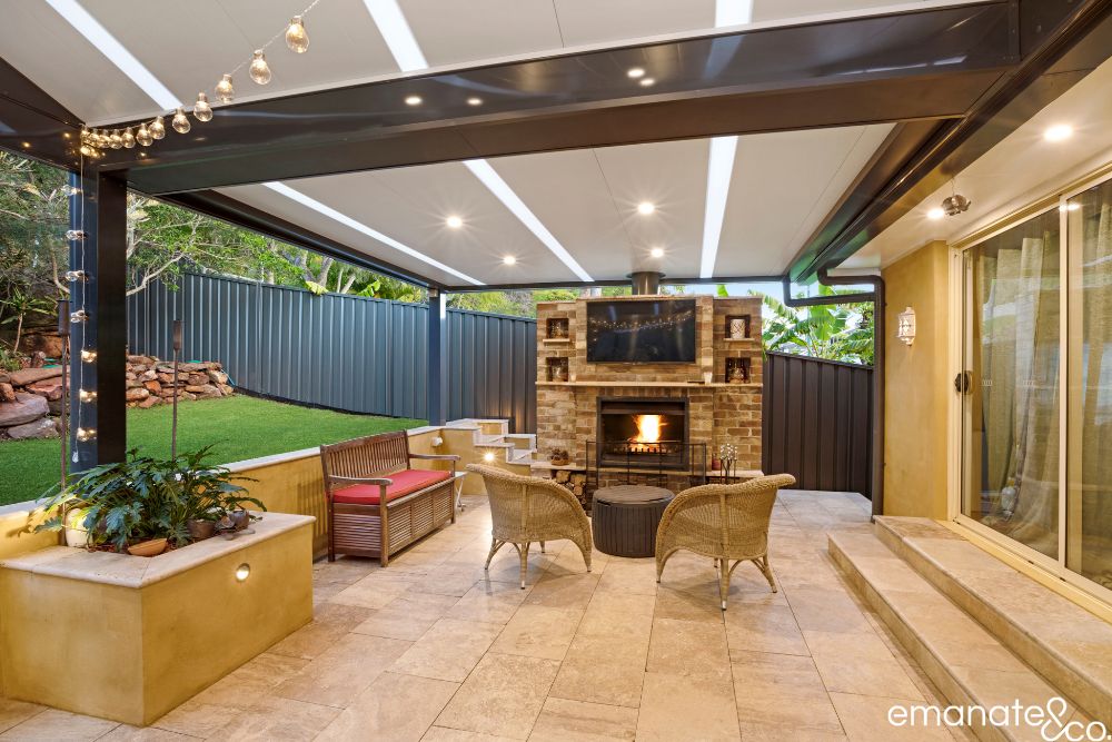 Stone-paved alfresco lounge beneath an insulated patio roof, featuring a brick fireplace, mounted television and integrated lighting at a Sydney home.